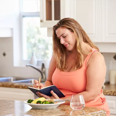 A woman eating a meal in her kitchen and tracking the meal in a journal