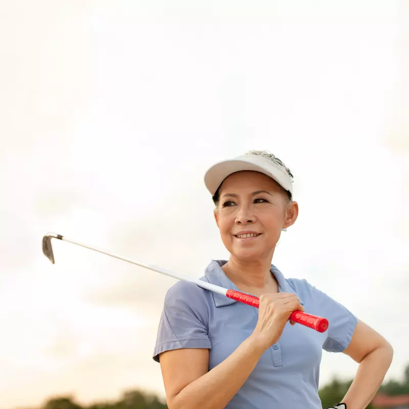A senior woman smiles while holding onto a golf club that is resting on her shoulder.